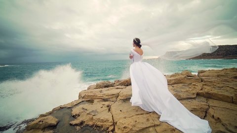 Amazing view a young bride in a white wedding dress and veil standing on a rock against the backdrop of sea or ocean and crashing waves Attractive young woman in nature on a background of the landscape.