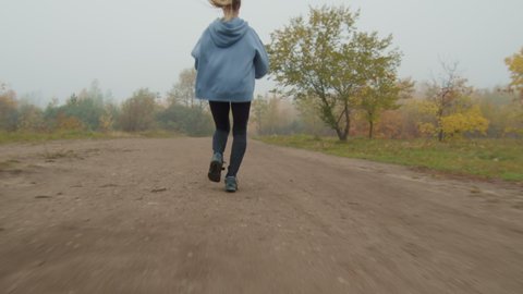 Slowmo tracking rear view of young woman in hoodie jogging along path in park on foggy autumn day