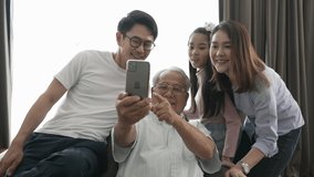 Happy Asian family sitting on sofa smiling old man holding smartphone taking selfie happily. Cheerful family smiling selfie enjoying together old man uncle holding smartphone talking video of family.