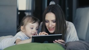 Happy Family Young Mum And Cute Little Kid Daughter Use Digital Tablet Technology Device Look At Screen, Mother With Child Surfing Internet Play Game Laying On Sofa At Home