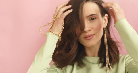 Lovely coquettish brunette woman, touching earrings, flirting. Woman fix her hair. Curly hair fashion model face smiling. Girl wear green dress, bracelet and long gold earrings on pink background.