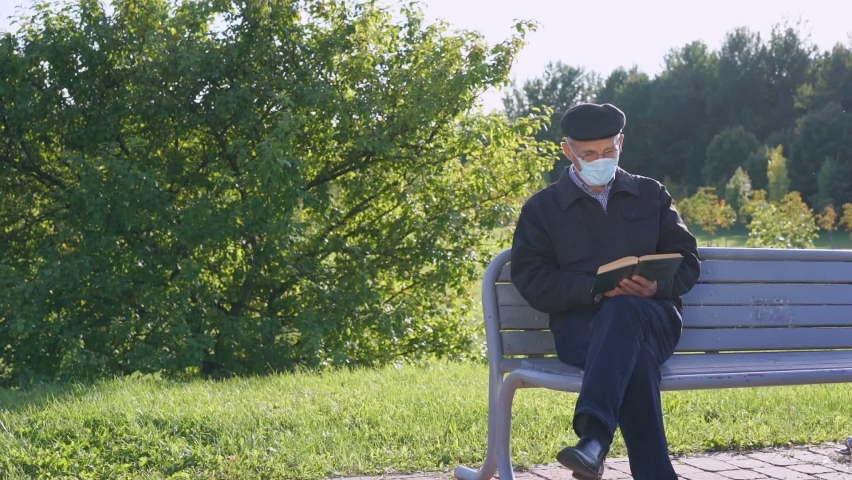 Elderly man in protective face mask sits on street bench and reads book.