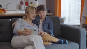 A little boy is watching funny video on his cell phone with his pregnant mother. Family, technology and pregnancy concept - happy pregnant mother and little son with smartphone at home.