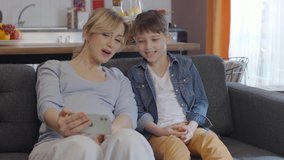 A little boy is watching funny video on his cell phone with his pregnant mother. Family, technology and pregnancy concept - happy pregnant mother and little son with smartphone at home.Slow motion.