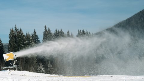 Snow cannon at ski resort in winter sun day. Snowmachine produced artificial snow at evergreen spruce forest, blue sky. Preparation of ski track in mountain active sport. Turbine sprays water in cold