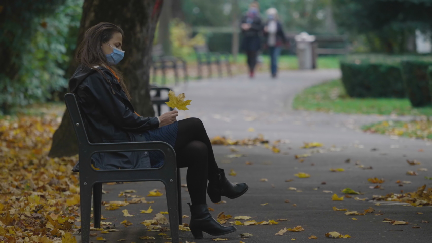 Single woman with protective mask sit on bench and play with autumn leave, enjoy