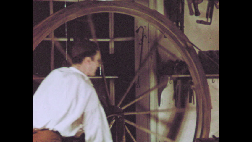 1960s: man spinning large wheel to rotate wooden rod while another man carves into rod, woman comes into store to look at items