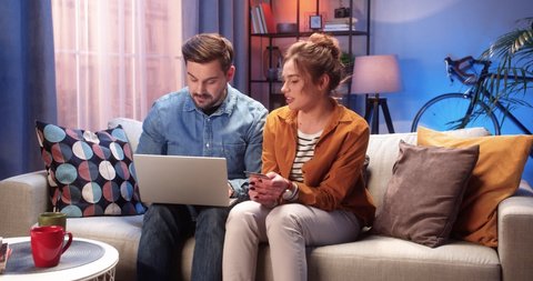 Happy young Caucasian married couple man and woman buying on internet on laptop making payment with credit card sitting on couch in house at night in good mood with satisfied faces, online shopping