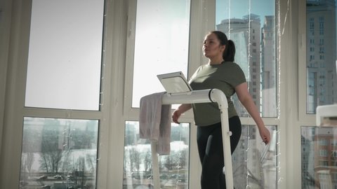 Young chubby woman is walking on treadmill in front of window. Motivated female is exercising on home smart trainer for weight loss, doing cardio fitness exercises. Sport activity. Healthy lifestyle
