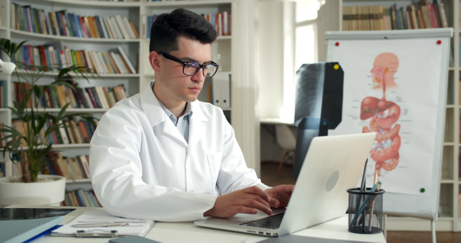 Male doctor opening laptop and started typing, inserting information from form. Young man in glasses and white rob working with personal computer while sitting in medical office.
