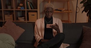 Crop view of serious millennial man watching video while sitting on sofa at home. Handsome afro american male person holding smartphone horizontally and looking at screen.