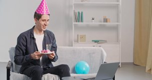 Caucasian lonely man sitting on sofa makes video call from home using laptop speaks into webcam accepts birthday greetings wears festive hat blows out candle on cake makes wish, party during lockdown