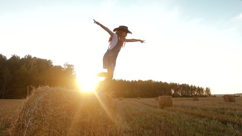 Young girl in denim overalls and a cowboy hat jumps off a bale of straw or hay and poses against the sunset. Child plays in a rural area in Sunny weather in the evening. Slow motion