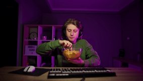 Portrait of a hungry gamer in casual clothes and headset having dinner at night at the computer with chips from a plate and closing his eyes with pleasure.