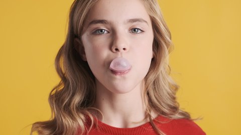 Close up happy blond teenager girl blowing bubble from chewing gum on camera over yellow background. Candy bubble