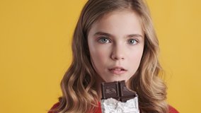 Close up blond teenager girl enjoying delicious chocolate bar on camera over yellow background. Very tasty expression