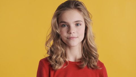 Beautiful blond teenager girl with wavy hair dressed in red sweater smiling on camera isolated on yellow background