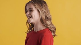 Pretty blond teenager girl in red sweater waving head and playing with hair fooling around on camera over yellow background. Cheerful emotion