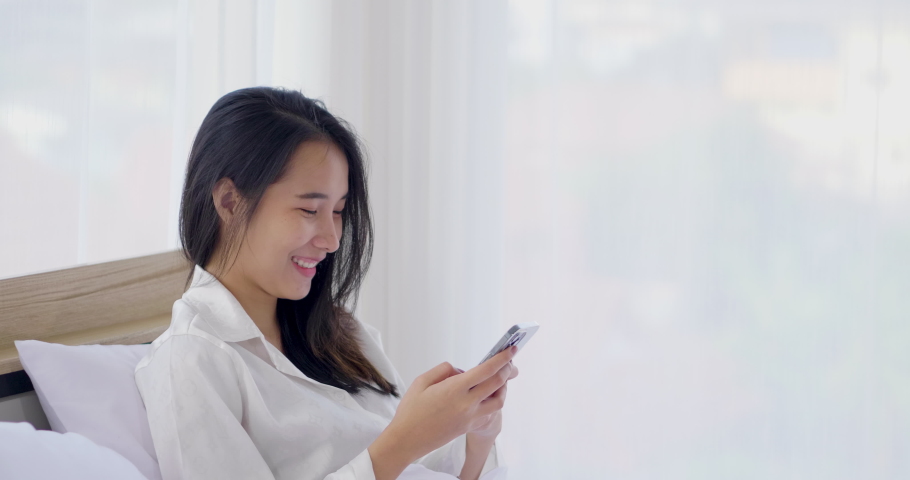 Young blogger Asian woman using smartphone social media to check rating response online page and writing to communicate with fan to keep relation. Relaxing chatting to friend on the bed in white shirt | Shutterstock HD Video #1068923153