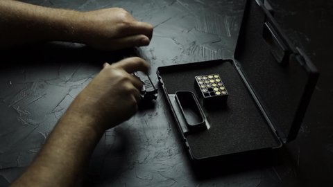 Men's hands open briefcase with firearms and ammunition, insert clip with bullets in gun and distort bolt. Preparing pistol for shooting on a black texture table.