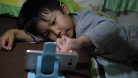 4K Asian child boy lying on bed, hand touch screen, watching video on smartphone, sneeze from dust allergy in room. Concept of technology, communication, health care.