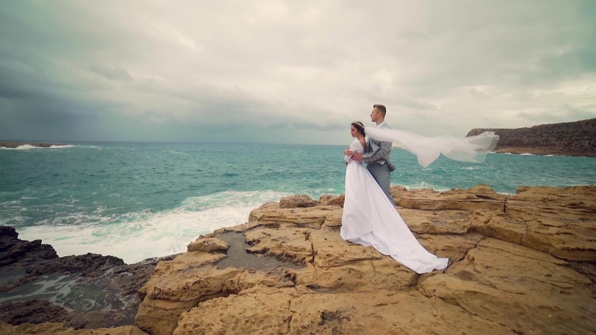 Wedding couple bride and groom stand a rock against backdrop ocean or sea hug together. Amazing view landscape of clouds and dramatic waves. white dress on a background of nature. wind blows the veil