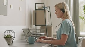 Medium close-up in profile of blond-haired Caucasian young woman sitting by desk at home office having video conference via laptop. Girl using earphones with mic, gesticulating, smiling, talking