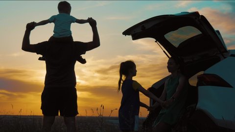 Silhouette the happy family of four people, mother, father and two children are happy sitting in the open trunk of a car at the sunset time. Concept of summer vacation and friendly family.