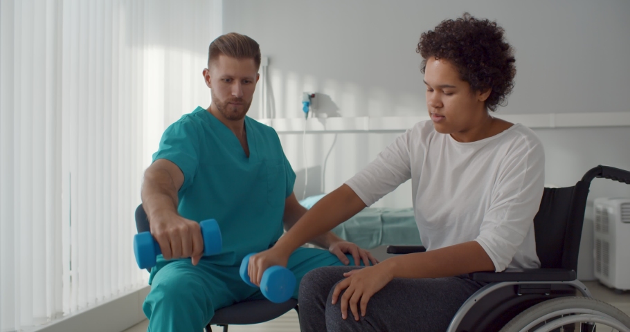 Doctor helping african woman in wheelchair outstretching arms with dumbbells during rehabilitation exercise in modern medical center. Disabled afro-american female having physiotherapy, Royalty-Free Stock Footage #1068930404