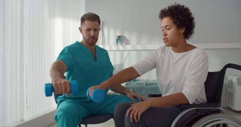 Doctor helping african woman in wheelchair outstretching arms with dumbbells during rehabilitation exercise in modern medical center. Disabled afro-american female having physiotherapy,