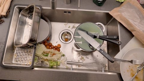 
A sink after cooking. Vegetables, unwashed plates and pots are in the sink. Camera moves towards the drain where leftovers are collected in a sink strainer.