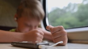 Closeup view video of blurry defocused cheerful happy little european boy traveling by train and listening to music looking in window. Focus at phone laying on white table in foreground