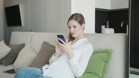Relaxed Young Woman Using Smart Phone Surfing Social Media, Checking News, Playing Mobile Games or Texting Messages Sitting On Sofa. Millennial Lady Spending Time At Home With Cell Gadget Technology