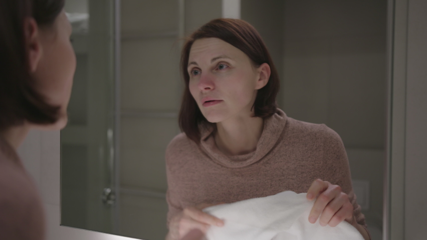 Young adult woman wiping her face with white towel looking at mirror in bathroom.