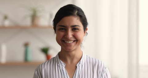 Head shot smiling pretty millennial indian ethnicity woman looking at camera, feeling confident indoors. Happy young multiracial lady posing alone at own home, showing healthy white toothy smile.