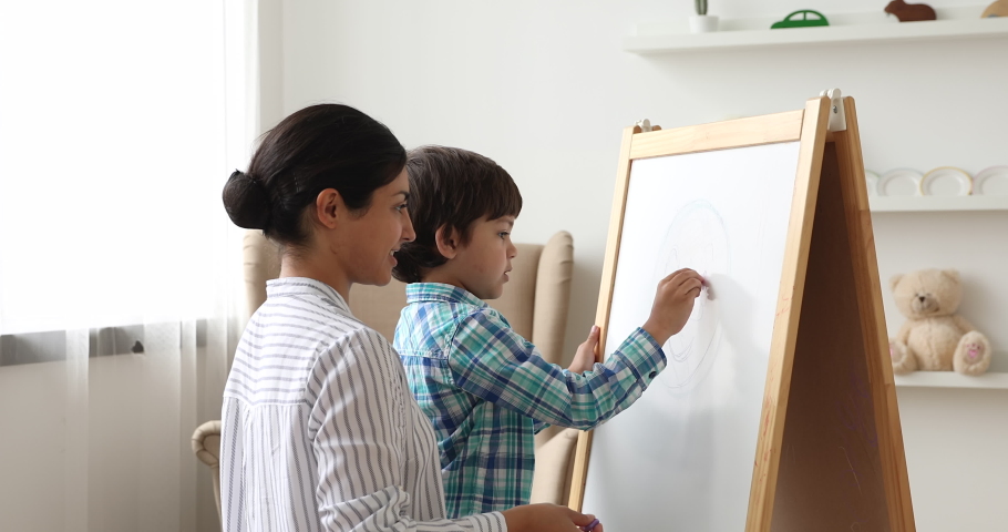 Affectionate young indian ethnicity mother helping or teaching small preschool cute child son drawing on white board with colored crayons. Happy family enjoying creative weekend pastime at home. Royalty-Free Stock Footage #1068950009