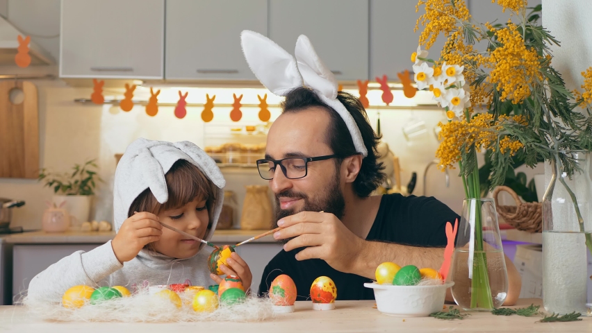 Happy Easter. Father in rabbit ears and young son draw on painted eggs at the table, at home. Funny family prepares for holiday Royalty-Free Stock Footage #1068950774
