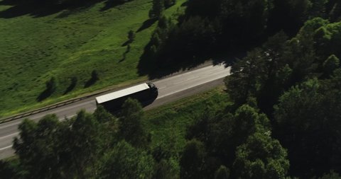 One Semi Truck with white trailer and black cab driving, traveling alone on dense flat forest asphalt straight road, highway top view follow vehicle aerial footage. Freeway trucks traffic