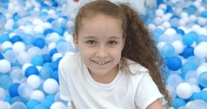 Portrait of a cheerful little girl, smiling child, jumps out of children's plastic balls,kid looks at the camera, a cute baby girl with a beautiful face play at home, rejoices. Happy childhood concept