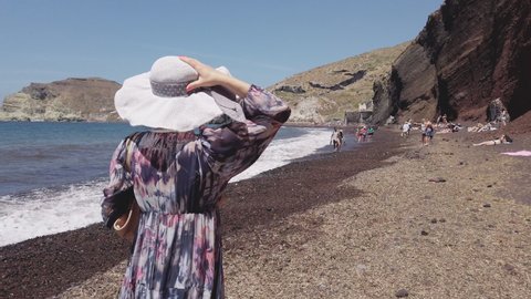 Santorini, Greece - 15 May, 2020: Tourist woman with hat walking on Red beach by Aegean sea in Akrotiri, Santorini island, slow motion