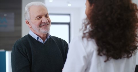 Happy elderly male patient comes up to female doctor shaking hands thanks for care and help in recovery, smiling each other standing in hospital. Mixed race nurse and gray haired caucasian man