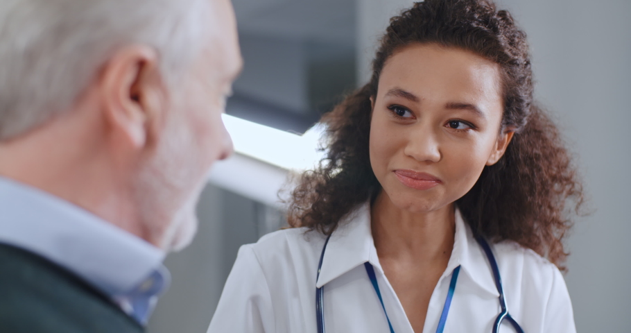 Portrait multiethnic female doctor in white medical coat and satisfied senior patient talk about good news from his health report and smiling in hospital. Mixed race nurse and elderly caucasian man Royalty-Free Stock Footage #1068958133