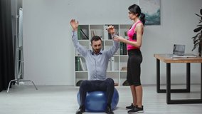 young fitness trainer helping businessman exercising on fitness ball in office