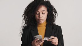 Young African American woman paying for online purchases by credit card using smartphone and rejoicing on camera over white background. Modern technology
