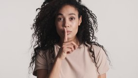 Young African American woman keeping fore finger over lips looking confident showing it is secret expression over white background. Do not tell it 