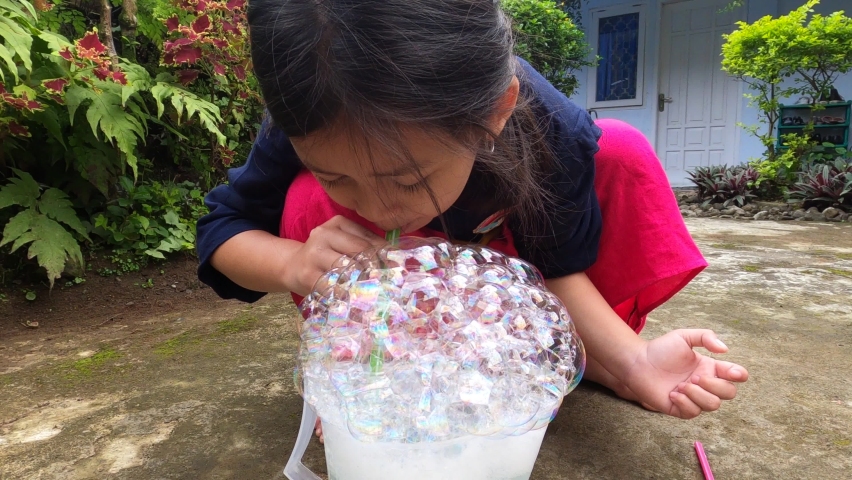 Adorable beautiful cute asian little girl enjoy playing blowing bubble soap using green straw with her mouth at the garden house yard with joy happy, fun during coronavirus covid pandemic new normal