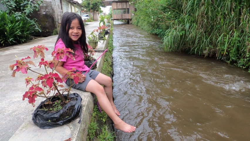 A pretty adorable beautiful cute asian little girl wearing pink shirt sit on edge of dirty brown small river enjoy playing water using foot with joy happy near street house neighborhood in daylight.