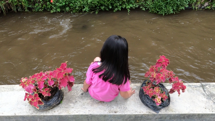 Pretty adorable beautiful cute asian little girl wearing pink shirt sit on edge of dirty brown small river enjoy playing water using foot with joy happy between tropical red leaves plants in daylight.