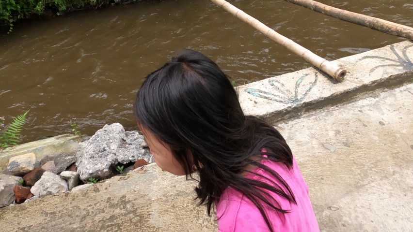 A pretty adorable beautiful cute asian little girl wearing pink shirt walking at the edge of dirty brown small river enjoy dancing with joy ugly weird happy near street house neighborhood in daylight.
