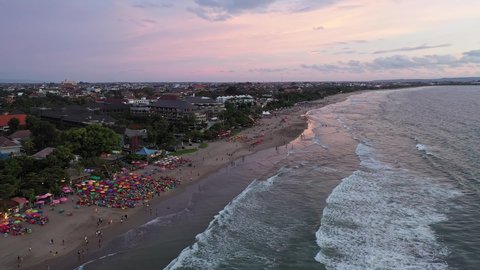 Aerial drone footage with backward motion of tourists enjoying a drink at a beach bar and restaurant on the famous Seminyak beach during sunset in Kuta, Bali, Indonesia. 